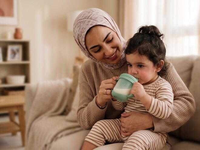 Photograph: smiling mother in hijab holding toddler drinking from cup on living-room couch