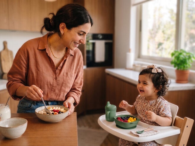 Photograph of mother smiling while feeding toddler in high chair in a sunny kitchen